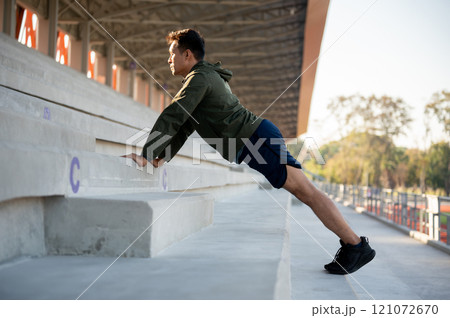A strong man in sportswear stretches his body with steps on a stadium, warming up before running. A strong man in sportswear stretches his body with steps on a stadium, warming up before running. 121072670