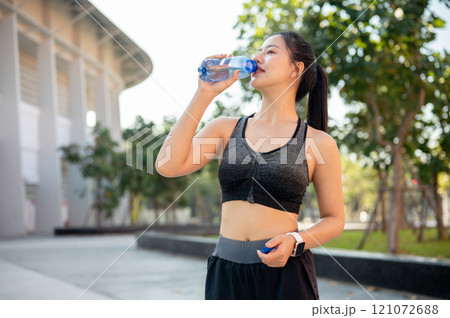 An attractive, healthy Asian woman in sportswear drinks water from a bottle after training outdoor. An attractive, healthy Asian woman in sportswear drinks water from a bottle after training outdoor. 121072688