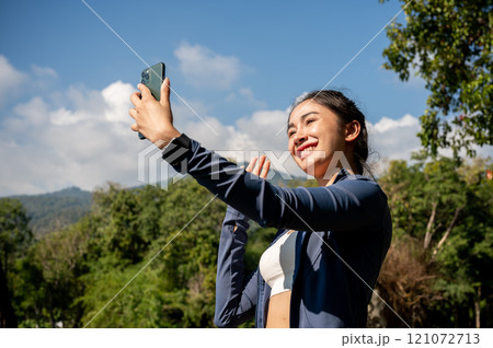 A cheerful Asian woman in sportswear takes a selfie on her smartphone while exercising in the park. 121072713