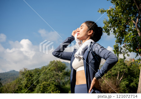 Tired, sweaty Asian woman in sportswear wipes sweat from her face with a towel, exercising in a park 121072714