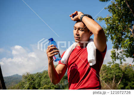 A sweaty, tired Asian man in sportswear drinks water while running in a park on a hot, sunny day. A sweaty, tired Asian man in sportswear drinks water while running in a park on a hot, sunny day. 121072726