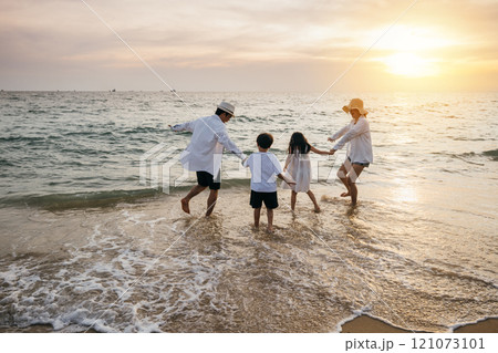 A carefree day at beach for an Asian family. Parents and daughters seen from behind enjoy bonding moments playing running and pretending to fly with arms outstretched. Family joy in sunlight. 121073101