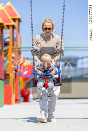 Mother pushing her infant baby boy child on a swing on playground outdoors Mother pushing her infant baby boy child on a swing on playground outdoors 121077721