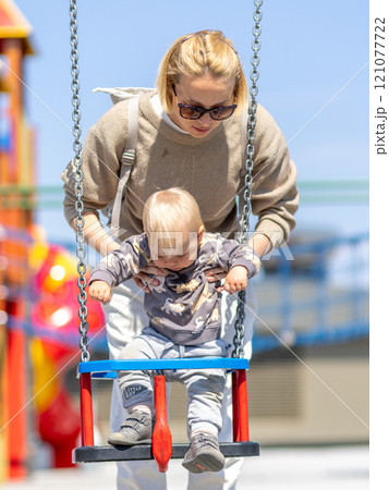 Mother pushing her infant baby boy child on a swing on playground outdoors Mother pushing her infant baby boy child on a swing on playground outdoors 121077722