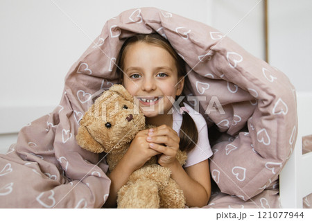Little Caucasian girl with missing teeth smiles while lying under blanket holding teddy bear. Brown heart-patterned quilt adds warmth and comfort. Happy childhood moment captured in bedroom setting. 121077944