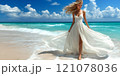 A beautiful female tourist in a light white dress walks along the beach. 121078036