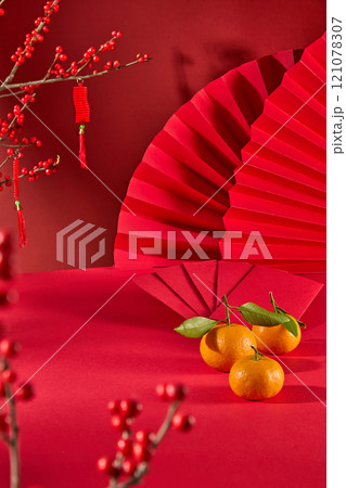 In a red backdrop, a close-up of an Asian New Year table with blank space display product is adorned with red envelopes, tangerines, a winterberry tree hanging fortunate strings, and red paper fans. 121078307