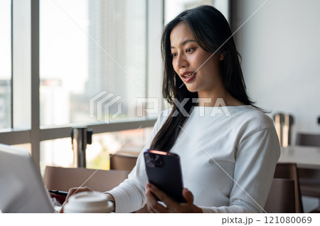 A woman holding her smartphone while working on her laptop computer, working in a modern office 121080069