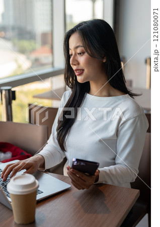 A woman holding her smartphone while working on her laptop computer, working in a modern office 121080071