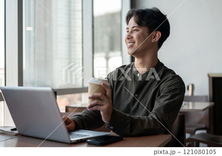 A positive Asian businessman enjoying a hot drink while working on his laptop, working remotely. 121080105
