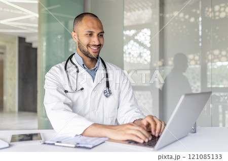 A smiling doctor is using a laptop in a modern office setting, showcasing productivity and professionalism. The image captures a sense of technology integration in the healthcare field. 121085133