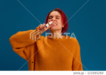 Portrait of young woman feels sick because of sneezing and spraying her nose to remove symptoms against dark blue background. 121085269