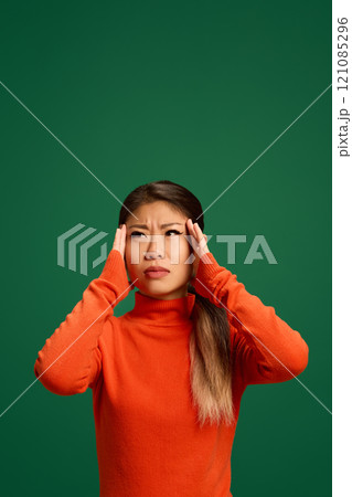 Young Asian woman holding hands on head as she feels pain dressed against green studio background. Woman feels badly. 121085296