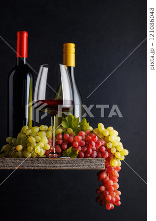 A glass and bottles of wine paired with grapes, set against a dark background A glass and bottles of wine paired with grapes, set against a dark background 121086048