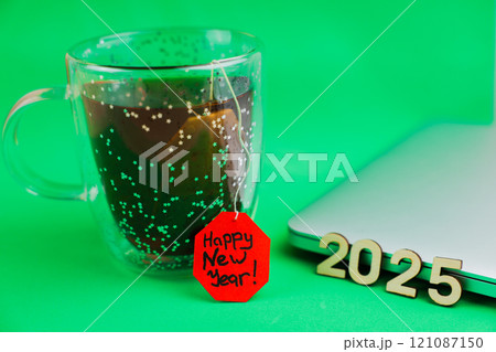 In the foreground of the photo on the side is a transparent mug with glitter with black tea in a bag, the label of which is bright red on which is written a happy New Year, next to it lies a closed In the foreground of the photo on the side is a transparent mug with glitter with black tea in a bag, the label of which is bright red on which is written a happy New Year, next to it lies a closed 121087150