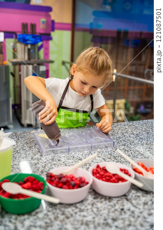 Little girl learning how to cook in a cooking class. Handmade dessert. Making chocolate candy, Child filling heart shaped mold with chocolate Little girl learning how to cook in a cooking class. Handmade dessert. Making chocolate candy, Child filling heart shaped mold with chocolate 121087501