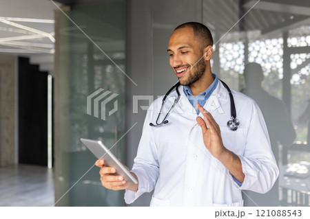 Professional doctor wearing stethoscope having friendly video call on tablet. Smiling and gesturing, showcasing modern healthcare communication and digital interaction in medical environment. 121088543