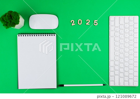 A white English computer keyboard flies to the right of the photo, on top are wooden numbers 2025, an office green plant and a white computer mouse, on the left lies a white notebook with a black 121089672