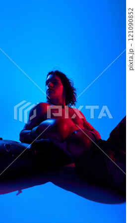 Handsome young shirtless man with muscular body. in boxing gloves resting punching bag against blue studio background in neon light Handsome young shirtless man with muscular body. in boxing gloves resting punching bag against blue studio background in neon light 121090852
