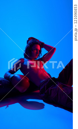 Handsome young shirtless man with muscular body. in boxing gloves resting punching bag against blue studio background in neon light 121090853