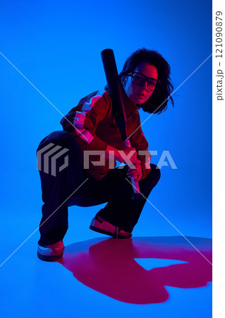 Stylish confident young woman in glasses and sportive attire, sitting and posing with baseball bat against blue studio background in neon light 121090879