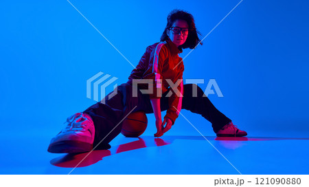 Stylish young woman in glasses and sportive jacket sitting with ball and posing against blue studio background in neon light 121090880