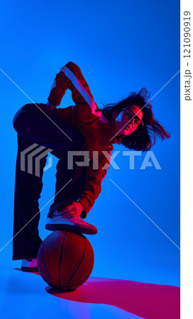 Young woman wearing red sports jacket and glasses posing dynamically with basketball against blue studio background in neon light 121090919