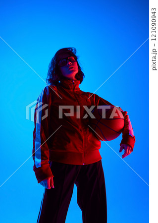 Portrait of beautiful young woman in sportswear standing in confidence pose and posing with basketball against blue studio background in neon light Portrait of beautiful young woman in sportswear standing in confidence pose and posing with basketball against blue studio background in neon light 121090943