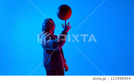 Handsome young guy in bomber jacket spinning basketball ball on finger against blue studio background in neon light 121090944