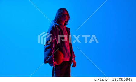 Side view portrait of handsome young man with long curly hair, wearing oversized sportive clothes standing with basketball against blue studio background in neon light Side view portrait of handsome young man with long curly hair, wearing oversized sportive clothes standing with basketball against blue studio background in neon light 121090945