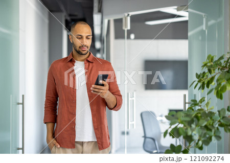 Young man in casual attire stands inside office using smartphone. Focused and connected, navigating workspace with phone in hand, exemplifying modern work style and communication. 121092274