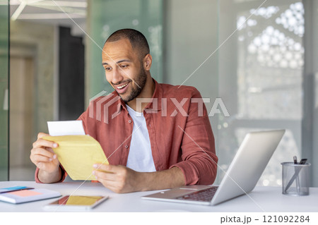 A cheerful man opening and reading an envelope at his office desk. He appears pleased with the contents, symbolizing success, important news, or professional correspondence in a lively workspace. A cheerful man opening and reading an envelope at his office desk. He appears pleased with the contents, symbolizing success, important news, or professional correspondence in a lively workspace. 121092284