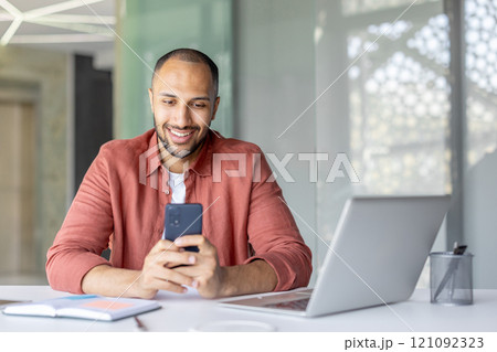 A cheerful male professional interacts with his smartphone while multitasking, working at his laptop. His positive demeanor embodies efficiency, communication, and productivity in work environment. 121092323