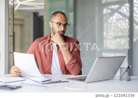 A focused professional in a modern office working with documents and a laptop computer, expressing concentration and analysis. A focused professional in a modern office working with documents and a laptop computer, expressing concentration and analysis. 121092336