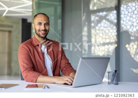 Smiling man working on laptop at desk in modern office, showcasing productivity. Table holds smartphone and pen holder, reflecting efficiency and work environment. Bright, professional setting. 121092363