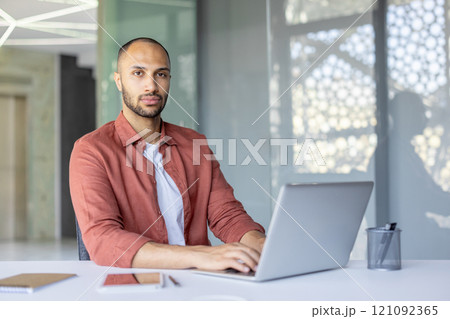 A professional man seated at a desk working on a laptop in a modern workspace, conveying focus and productivity. Includes office supplies and a bright, open environment. 121092365