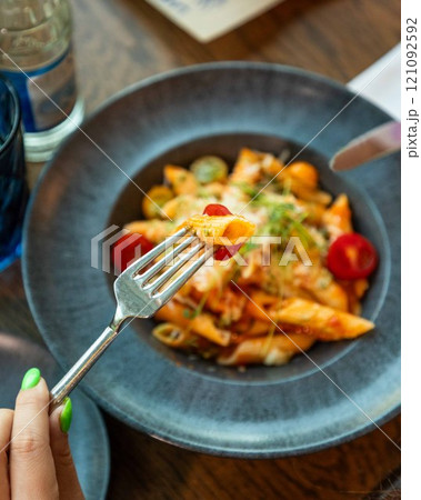 Woman holding penne pasta on fork over plate of food Woman holding penne pasta on fork over plate of food 121092592