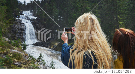 Woman, friends and camera with waterfall for sightseeing, picture or photography in nature. Female person, tourist or traveler with phone screen for memory, view or adventure by river in forest 121094298