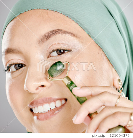 Muslim woman smiling and using a facial roller on her skin. Beauty model doing her morning skin care routine with a jade roller. Young female using a gua sha product for healthy smooth skin. 121094373