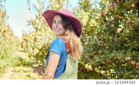 Portrait of a beautiful female farm worker standing on a fruit farm during harvest season. Young happy farmer between fruit trees on a sunny day in summer. Agricultural industry growing fresh produce 121094395