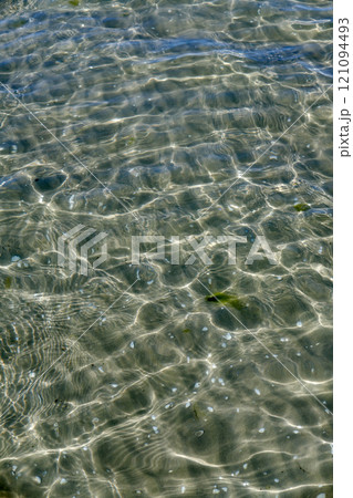 A clear water surface on the sea coast with the seashell and wavy water on a sunny day. The top view of transparent sea water at the seashore with the reflection of scattered sunlight under the water 121094493