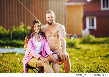 White shirtless man and woman in shorts pose for photographer on lawn near house, seated portrait of married couple in their 20s. 121096914
