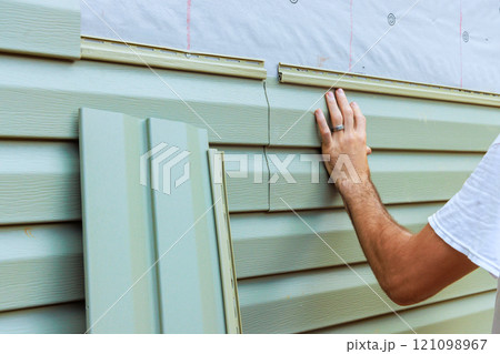 Construction worker carefully aligns, secures new vinyl siding panels on to exterior wall of new house Construction worker carefully aligns, secures new vinyl siding panels on to exterior wall of new house 121098967