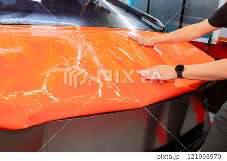Person is smoothing out an orange vinyl wrap on car hood in workshop, ensuring bubble free finish for protect body automotive 121098970