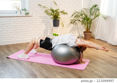 Young woman exercising on a stability ball in a bright room with plants Young woman exercising on a stability ball in a bright room with plants 121099997