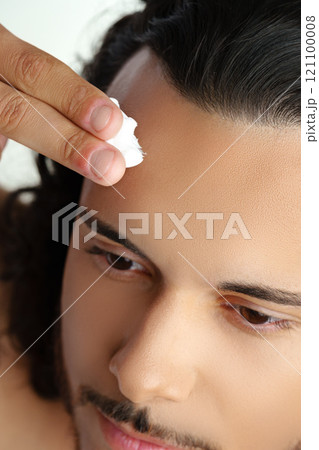 A young man applying skincare cream on his face with a focused expression, demonstrating personal grooming at home 121100008