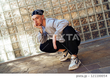 A young man wearing a gray sweatshirt and black pants squats in front of a glass block wall A young man wearing a gray sweatshirt and black pants squats in front of a glass block wall 121100054