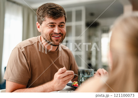 A man smiles while eating with a friend at a dining table 121100073