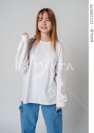 A joyful young girl with long hair wearing a white sweater stands against a plain background 121100079
