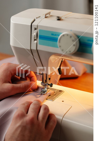 Hands sewing fabric using a sewing machine on a wooden table in a well-lit workspace Hands sewing fabric using a sewing machine on a wooden table in a well-lit workspace 121100141